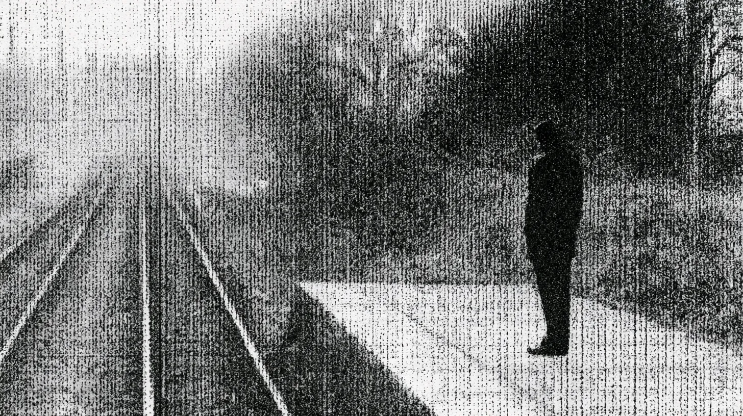 A lone figure standing on a railway platform, silhouetted against the tracks and trees beyond.