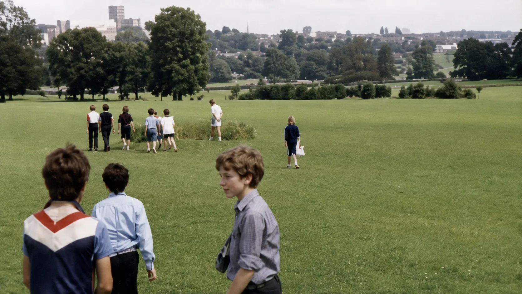 Schoolchildren in a park in the mid-1980s, several walking away across mown grass towards trees, with a town and tower blocks on the skyline behind.