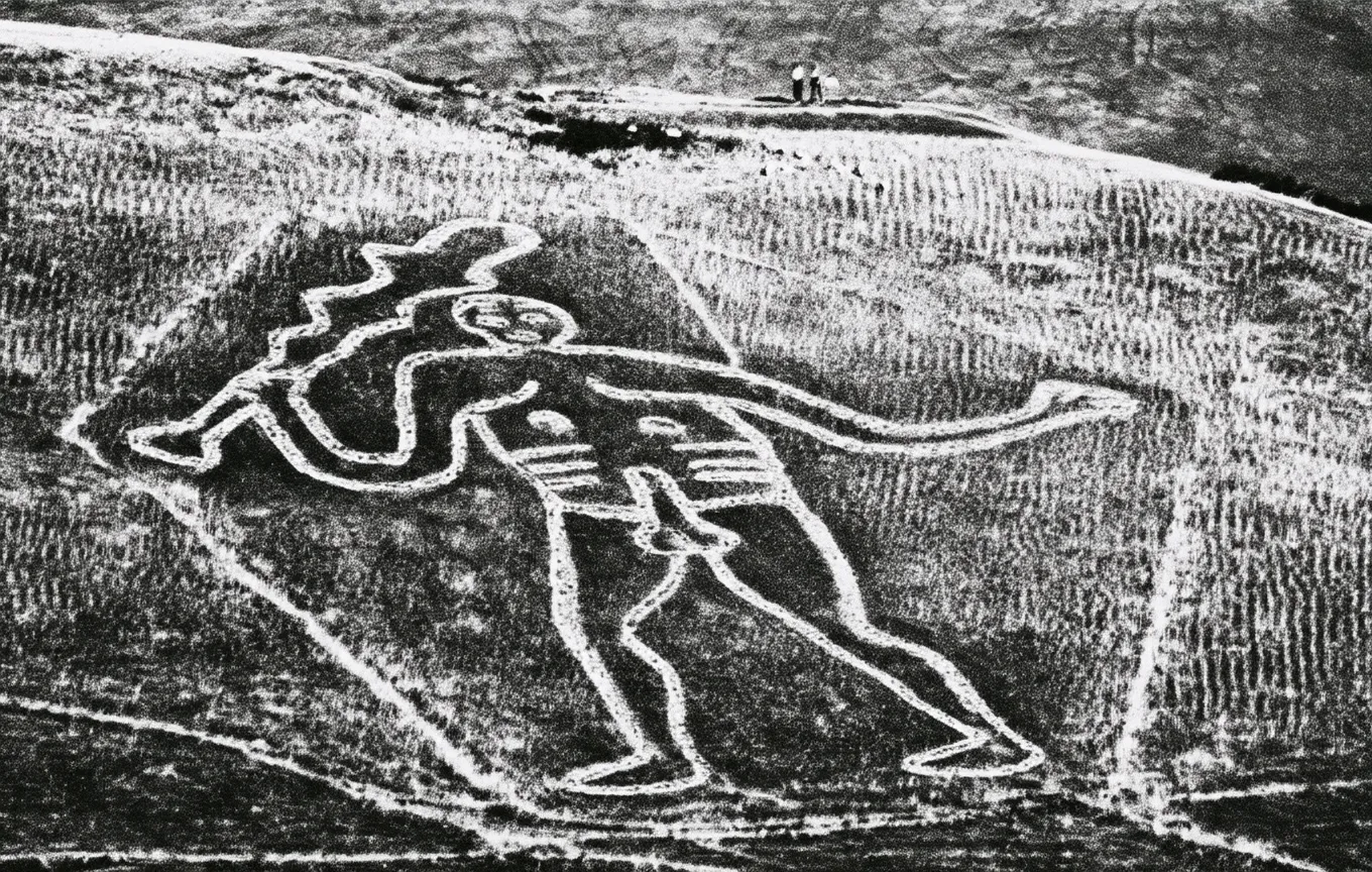 An aerial view of the Cerne Abbas Giant hill figure cut into a chalk hillside, two onlookers standing at its head.