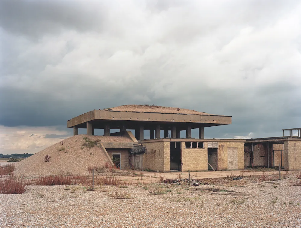 The concrete slab roof of one of the Orford Ness pagodas, raised on stubby columns above a shingle bank, derelict brick outbuildings alongside it under a heavy grey sky.