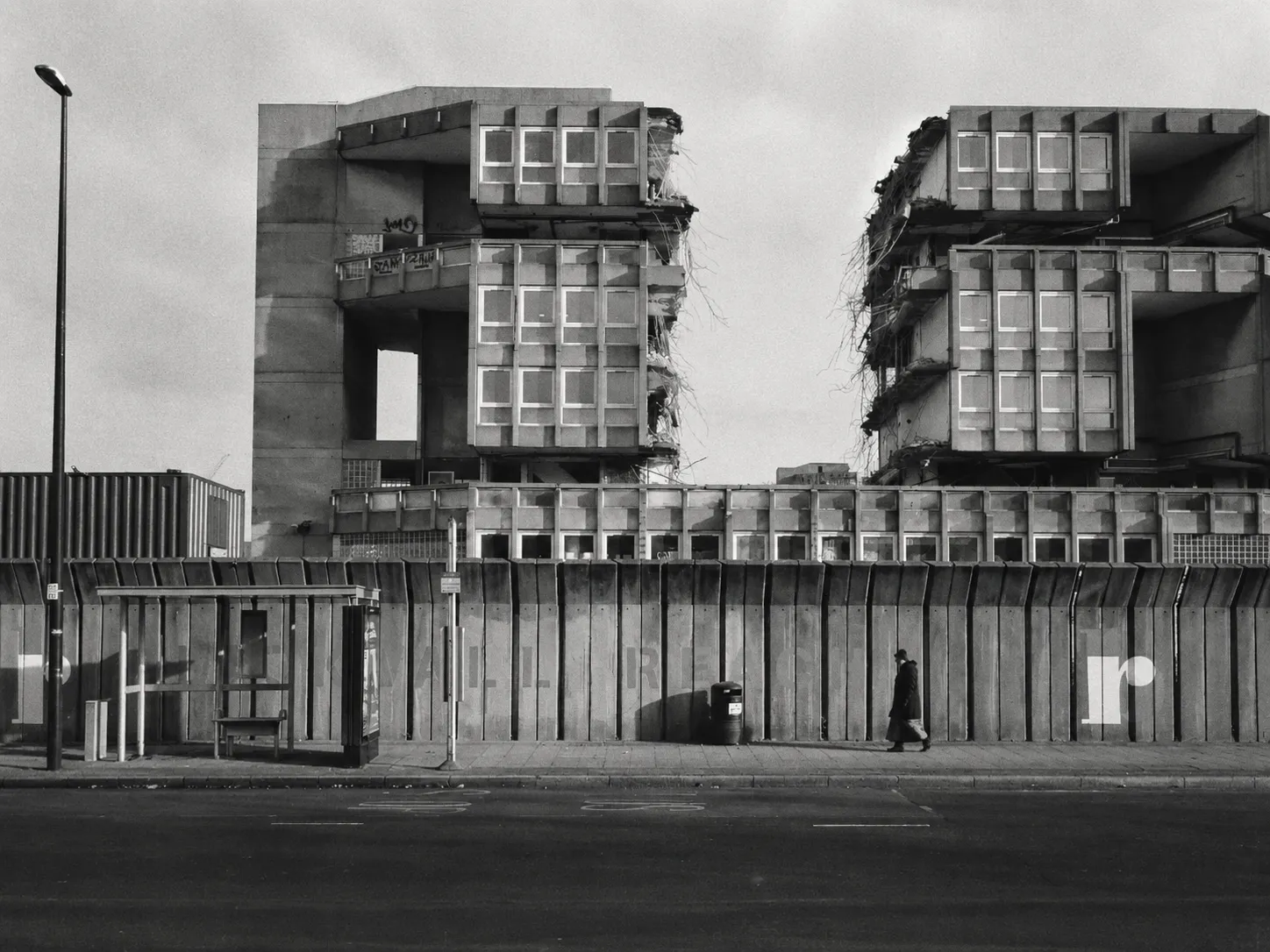 Robin Hood Gardens mid-demolition in black and white: two concrete slab blocks with their flank walls torn open, seen from across a road behind a line of hoarding, a bus shelter at one side and a single figure walking past.